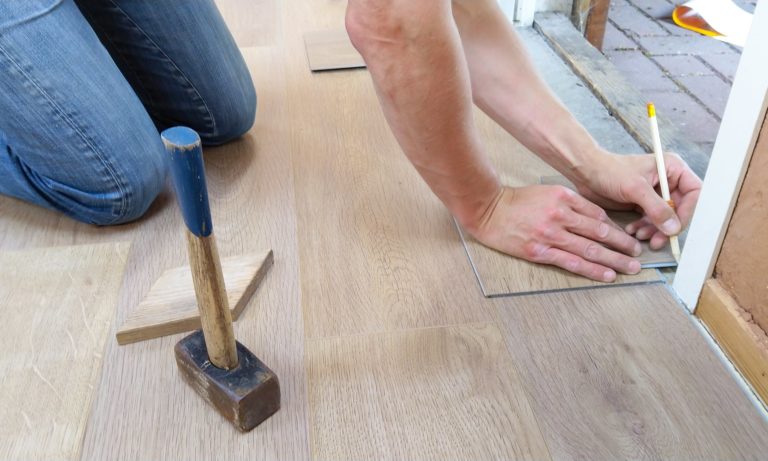 hands and knees of carpenter are visible as they measure door frame for flooring. pencil is in hand and hammer is nearby.