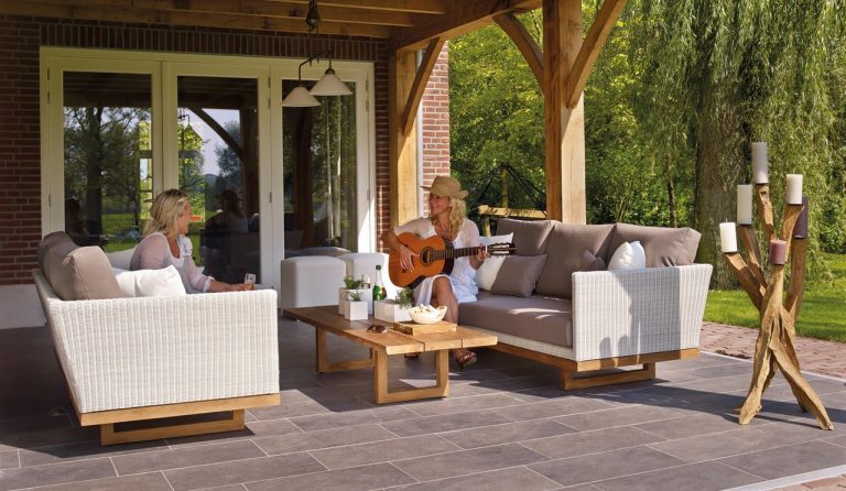 two smiling women lightly dressed sit on outdoor patio of home. one glays guitar. prominent in image is driftwood candleabra