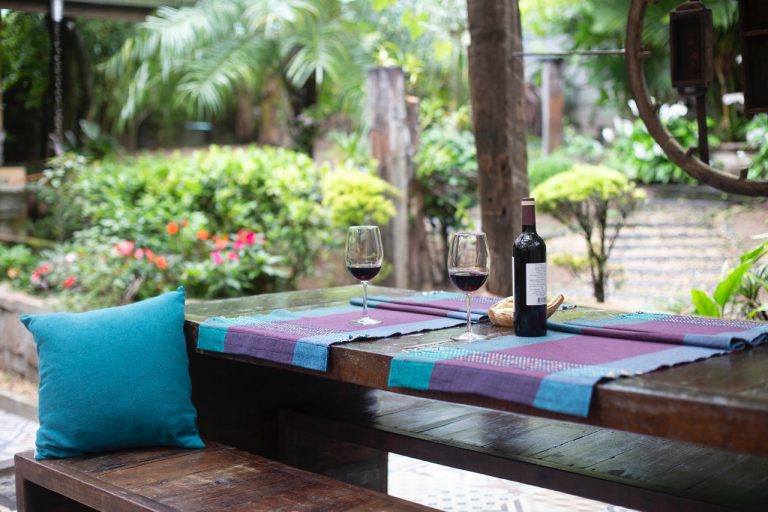 outdoors in a garden two glasses and a bottle of red wine on a dark wood table with blue and purple tablecloth and blue pillows