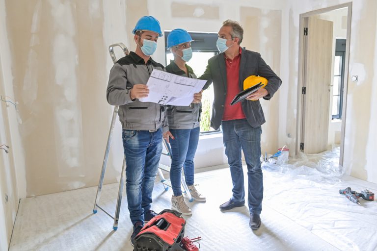 A home builder talking to two construction workers during a home renovation. All are wearing blue medical masks.