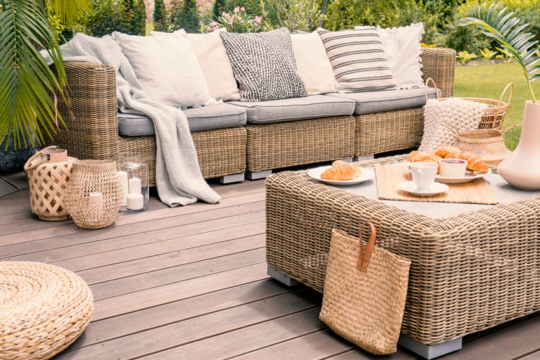brown outdoor wicker patio set with grey and white pillows. white tea set with croissants. green ferns in background.