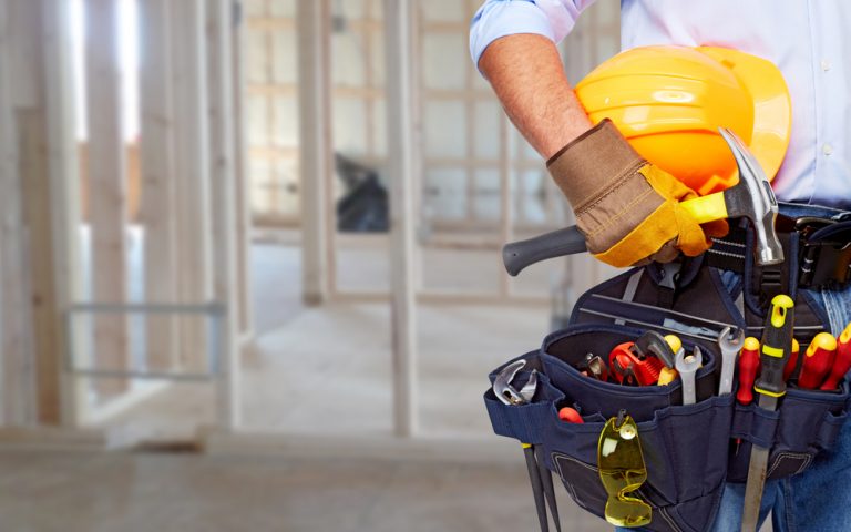 person with blue toolbelt holding hammer and yellow construction helmet. out of focus house frame in background.