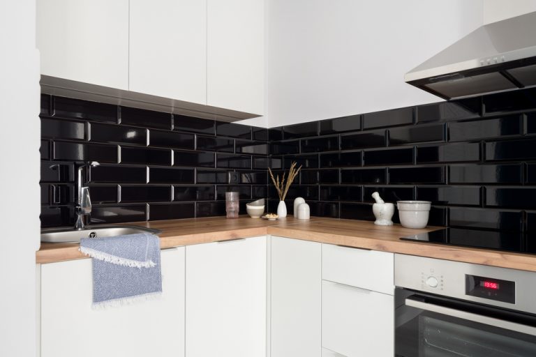 Classic and simple designed kitchen with white furniture, black backsplash tiles, wooden countertop and silver oven, kitchen hood and sink with tap